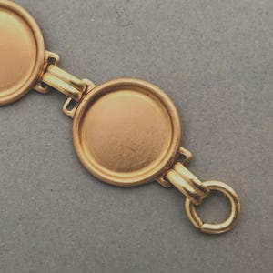 May include: A gold-toned metal bracelet composed of three circular, flat-surfaced bezel settings linked together by double bars, with a small circular jump ring attached to the end of the final setting.
