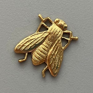 Op de afbeelding: Een gouden metalen vlieg-bedel met gedetailleerde vleugels en lichaam. De vlieg is driedimensionaal ontworpen en heeft een textuur.