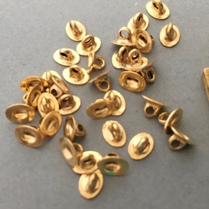 Puede incluir: Un conjunto de pequeños cierres metálicos dorados. Cada cierre tiene una base ovalada plana y un lazo para sujetar a la tela. Los cierres están dispersos sobre una superficie gris, posiblemente para un proyecto de costura.