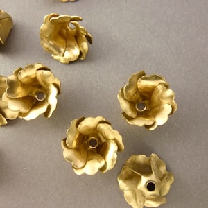 Puede incluir: Un conjunto de siete adornos de metal dorado en forma de flor con un agujero en el centro. Los adornos están dispuestos en semicírculo sobre una superficie gris.