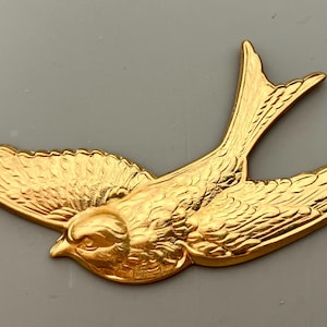 May include: A gold-colored metal bird figurine with detailed wings and a delicate beak. The bird is in flight, with its wings spread wide.