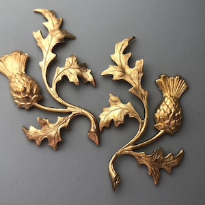May include: Two gold-toned metal thistle designs with intricate details. Each thistle has a stem and leaves, and the top of each thistle is a flower with a spiky center.