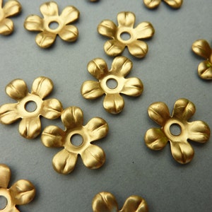 Puede incluir: Un grupo de adornos metálicos de color dorado en forma de flor con un agujero en el centro. Los adornos están esparcidos sobre una superficie gris.