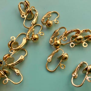 May include: A collection of gold-toned metal clip-on earring findings. Each piece features a hinged mechanism with a small circular loop at the base for attaching charms or dangles. The components are scattered across the frame, showing the metallic finish.