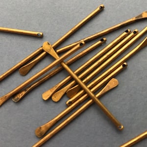 Puede incluir: Primer plano de numerosas y pequeñas agujas de metal de color latón antiguo. Cada aguja tiene un extremo plano en forma de paleta y un pequeño agujero en el extremo opuesto. Las agujas están dispersas sobre una superficie gris.