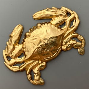 May include: A gold-colored metal crab ornament with detailed sculpting. The crab is in a curled position with its claws raised.