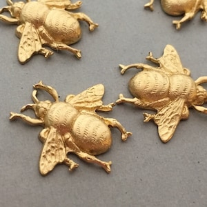 Pode incluir: Quatro pingentes de abelha de metal dourado com asas e corpos detalhados. As abelhas estão dispostas de forma dispersa sobre uma superfície cinza.