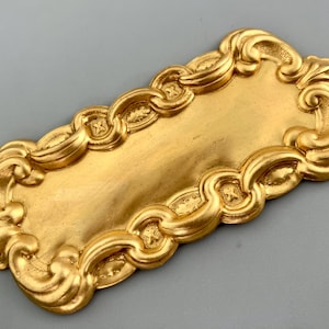 May include: A rectangular gold-colored metal plaque featuring a smooth, flat center panel surrounded by a raised, ornate border with repeating scrollwork and small cross-shaped details. The edges are scalloped with decorative flourishes.