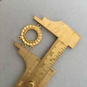 May include: Gold-toned metal pendant with a circular design and three jump rings. The pendant is being measured with a ruler, showing it is approximately 1 inch in diameter.