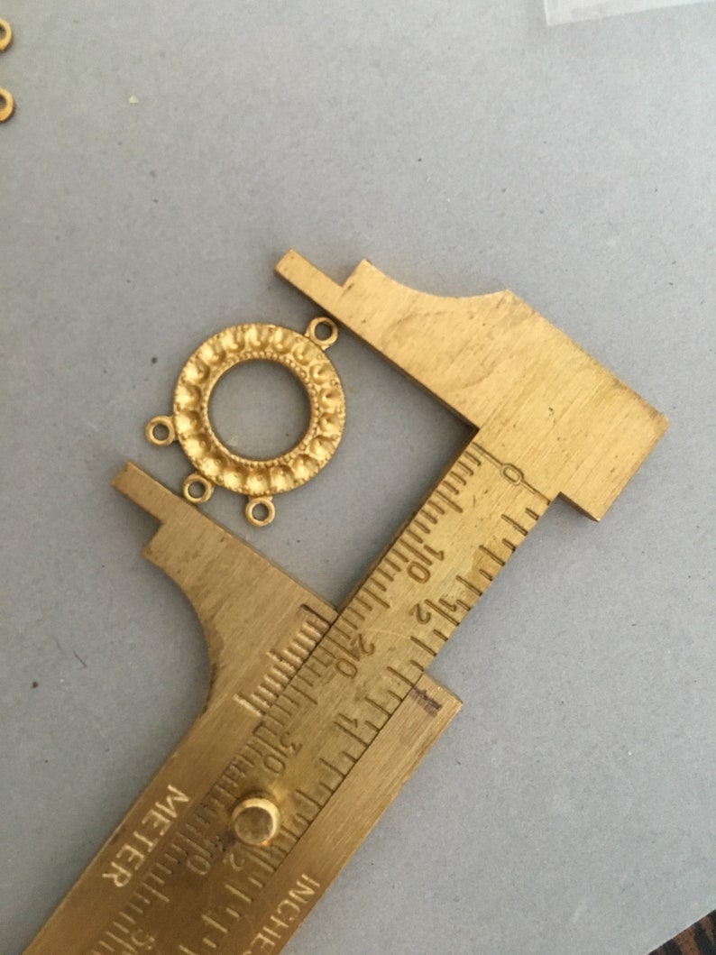 May include: Gold-toned metal pendant with a circular design and three jump rings. The pendant is being measured with a ruler, showing it is approximately 1 inch in diameter.