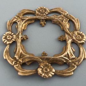 Puede incluir: Un marco cuadrado de latón con un diseño calado ornamentado, cuatro motivos florales en cada lado y elementos metálicos curvos que conectan las esquinas. El metal tiene un acabado dorado cálido con una textura visible.