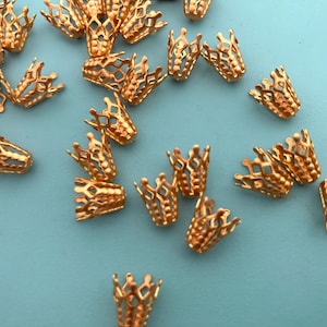 May include: A collection of small, copper-colored metal bead caps featuring a delicate, openwork filigree crown design. Each piece has a scalloped top edge and a tapered, cylindrical shape, scattered across a solid light blue surface.