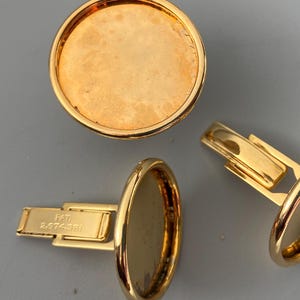 Può includere: Un paio di gemelli in oro con una superficie rotonda, liscia e dorata. I gemelli hanno una chiusura a cerniera con il testo "PAT. 2,874,381" inciso su uno dei fermagli.