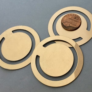 May include: Three gold-colored metal circle bookmarks with a slot cut out of the outer ring. One bookmark has a penny on top. The bookmarks are on a gray background.