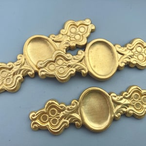 May include: Two gold-colored metal furniture pulls featuring an elongated, ornate design with a central oval recessed area and decorative scrollwork patterns on both sides. The hardware has a matte finish and symmetrical detailing.