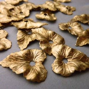 Puede incluir: Una colección de abalorios de metal de color dorado en forma de hoja con un agujero en el centro. Los abalorios están esparcidos sobre una superficie gris.