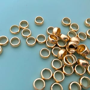May include: A collection of small, gold-colored metal rings scattered on a light blue surface. The rings are round with a smooth, polished finish, and appear to be jewelry components or craft supplies.