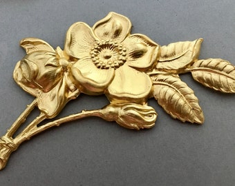 May include: A gold-colored metal flower with leaves and a bud. The flower has five petals and a center with a raised design. The leaves are detailed and realistic.