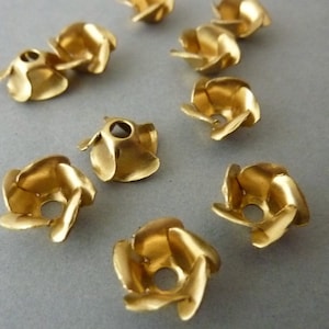 May include: Close-up of several small, gold-colored metal flower-shaped bead caps. Each cap has a central hole and curved petals. The caps are scattered on a neutral gray surface, ideal for jewelry making.