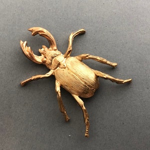 May include: A gold-colored beetle figurine with detailed texture. The beetle has large mandibles and six legs, with a segmented body. The figurine is set against a gray background.