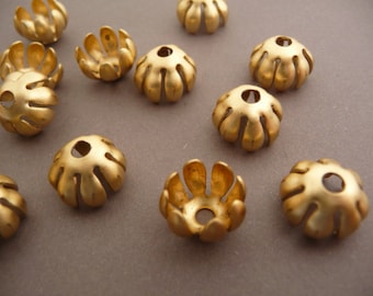 May include: Close-up of several gold-colored bead caps. Each cap has a flower-like design with multiple petal-shaped cutouts. The caps are scattered on a neutral gray surface, showcasing their intricate details and metallic sheen. These are ideal for jewelry making.