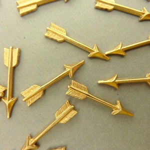 Könnte beinhalten: Eine Streuung von goldfarbenen Pfeil-Anhängern, jeder mit einer spitzen Spitze und einem dekorativen Federdesign. Diese Anhänger sind perfekt, um Schmuck, Kunsthandwerk oder Wohnaccessoires einen Hauch von Verspieltheit zu verleihen.