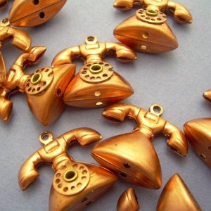 Pode incluir: Uma coleção de pingentes de telefone cor de cobre. Cada pingente apresenta um design clássico de telefone rotativo com um fone, um mostrador e um pequeno laço para prender a joias ou artesanato. Os pingentes têm um acabamento metálico.