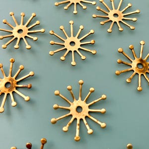 May include: Close-up of several brass-colored starburst-shaped decorative elements. Each starburst has a central hole and multiple radiating arms ending in small, round spheres. The objects are arranged against a muted teal background.