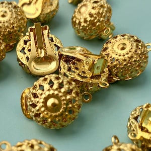 Peut inclure: Collection de boucles d'oreilles à clip dorées. Chaque boucle d'oreille présente un motif ajouré de forme ronde et un mécanisme à clip. Les boucles d'oreilles sont éparpillées sur une surface bleu clair.