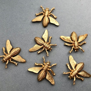 May include: Six gold-colored metal bee figurines with intricate details. The bees are arranged in a circular pattern on a gray background.