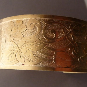 May include: A gold-toned metal cuff bracelet with an intricate floral design. The design features leaves and flowers, all etched into the metal.