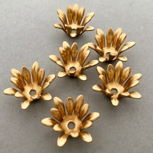 May include: Six gold-colored metal flower-shaped embellishments with a hole in the center. The embellishments have a scalloped edge and are perfect for adding a touch of elegance to crafts.