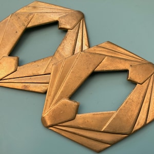 May include: Two Art Deco style brass-toned metal decorative hardware pieces. Each piece has a geometric design with a central opening and radiating lines. The hardware is likely for furniture or doors.