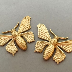 Könnte beinhalten: Zwei goldfarbene, mottenförmige Verzierungen. Jede Motte hat detaillierte Flügel und einen segmentierten Körper. Die Flügel sind ausgebreitet und die Antennen sind sichtbar. Die Verzierungen sind wahrscheinlich für Bastelarbeiten oder Schmuck.