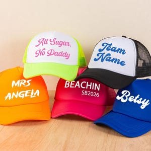 May include: Five trucker caps in orange, neon yellow, black, hot pink, and blue. The orange cap features MRS ANGELA, the yellow one reads All Sugar. No Daddy, the black one says Team Name, the pink one reads BEACHIN SB2026, and the blue one says Betsy.