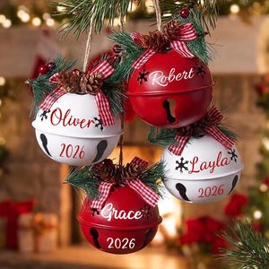 May include: Four round metal jingle bell ornaments hanging from pine branches. Two are white with red text reading Oliver and 2026 or Layla and 2026. Two are red with white text reading Robert or Grace and 2026. Each features a red plaid bow and pinecones.