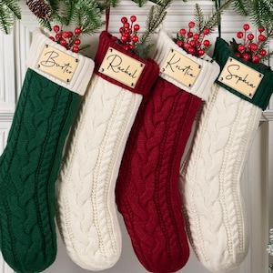 Op de afbeelding: Vier kabelgebreide kerstsokken in groen, wit en rood hangen aan een versierde schoorsteenmantel. Elke sok heeft een houten naamlabel. De namen op de labels zijn: Butler, Rachel, Kristin en Sophia. De sokken zijn versierd met takjes rode bessen.