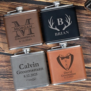Könnte beinhalten: Vier personalisierte Flachmänner mit silbernen Deckeln und lederartigen Oberflächen. Die Flachmänner sind braun, schwarz, grau und hellbraun. Jeder Flachmann hat einen Namen und ein Datum eingraviert. Der braune Flachmann trägt die Aufschrift "Morgan Groomsman 8.12.2025".