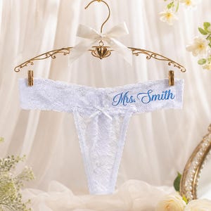 May include: A white lace thong featuring a small centre bow and the text Mrs. Smith embroidered in blue script. The garment is suspended from a gold-toned metal hanger adorned with a white satin ribbon bow at the top centre hook.