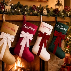 May include: Four Christmas stockings hanging in a row. The first is tan with a white bow and the name Nicola. The second is deep red with a white bow and the name Sophie. The third is white with a red bow and the name Henry. The fourth is dark green with a red bow and the name Willow.