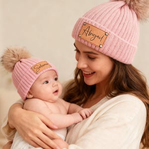 May include: Two pink knitted beanies with faux fur pom-poms. One beanie has a leather patch with the name "Abigail", and the other has "Salome". The hats are worn by a mother and baby.