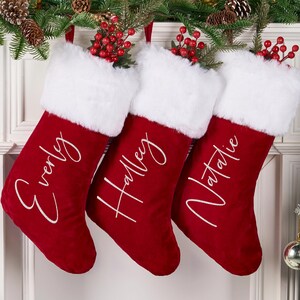Op de afbeelding: Drie rode kerstsokken met witte imitatiebont afwerking en gepersonaliseerde namen in cursief schrift. De sokken zijn versierd met takjes rode bessen en hangen aan een slinger van dennen en dennenappels.