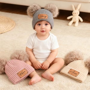 Könnte beinhalten: Ein Baby trägt eine graue Strickmütze mit zwei Kunstfell-Bommeln und einem Kunstlederetikett mit der Aufschrift "Henry". Zwei weitere Mützen, eine rosa und eine cremefarbene, befinden sich in der Nähe. Das Baby trägt einen weißen Body.