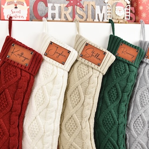May include: Five cable-knit Christmas stockings in red, cream, beige, green, and grey. Each stocking has a leather patch with a name: Kathryn, Jaylee, Stella, Ramsey, and Elanie. The stockings are hanging on a white shelf.