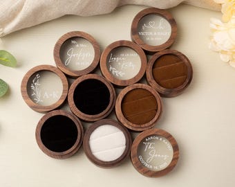 May include: A collection of round wooden ring boxes with clear lids. Some boxes have names and dates etched on them, while others have black, brown, or white velvet interiors. The boxes are arranged on a light-colored surface.