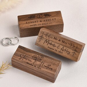 Puede incluir: Tres cajas de anillos de madera grabadas con nombres y fechas. Las cajas son de madera oscura y tienen un acabado natural. La caja superior tiene el texto "ROBERT & ASHLEY 08.15.2024-", la caja del medio tiene el texto "Mr & Mrs Michelle & Ashley August 26, 2024", y la caja inferior tiene el texto "Steven & Heather October 26, 2024".