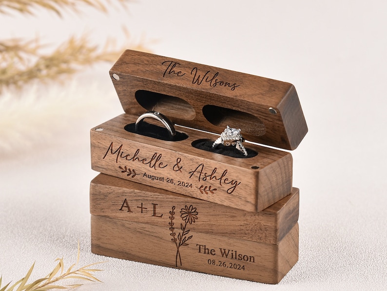 Puede incluir: Una caja de anillos de madera con dos compartimentos para anillos. La caja est&aacute; grabada con los nombres "Michelle & Ashley", la fecha "August 26, 2024", y el texto "The Wilsons".