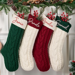 May include: Four cable-knit Christmas stockings in green, cream, and red hang from a garland. Each stocking has a white cuff with a name embroidered in cursive: Jamie, Pamela, Amelia, and Samuel. Red berries and greenery adorn the top.