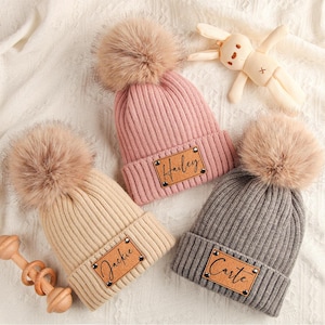 Peut inclure: Trois bonnets en tricot avec des pompons en fausse fourrure. L'un est rose, l'autre beige et le dernier gris. Chaque bonnet porte une étiquette en similicuir marron avec un nom. Un lapin en peluche et un jouet en bois sont également dans l'image.