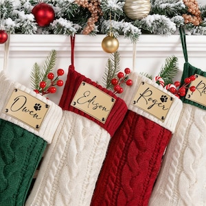 Könnte beinhalten: Vier personalisierte Weihnachtsstrümpfe aus grünem, rotem und weißem Zopfstrick. Jeder Strumpf hat ein Namensschild aus Holz mit einem Namen und einem Pfotenabdruck. Die Strümpfe sind mit Grün und Beeren geschmückt.